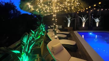 Zona de piscina con sillones reclinables, una sombrilla con luces de cadena y un elemento acuático.
