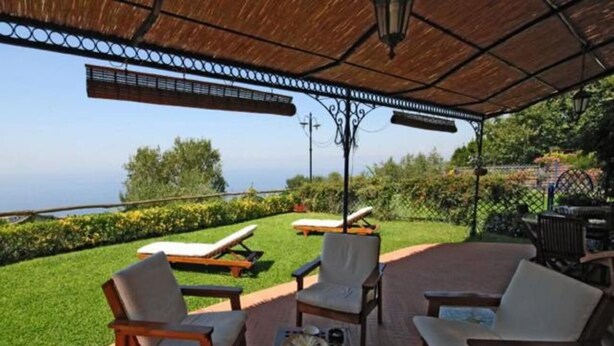 A covered outdoor seating area with lounge chairs and a table, overlooking a well-maintained lawn and a distant view.