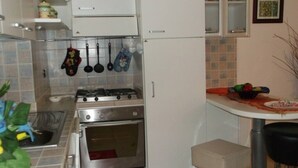 Eine Küche mit Gasherd, Backofen und hängenden Küchenutensilien. Ein Tisch mit einer Obstschale und ein Stuhl stehen bereit.