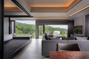 Una sala moderna con un amplio sofá, una mesa de centro de madera y un televisor. La habitación cuenta con ventanas grandes que ofrecen vistas a un paisaje verde y edificios.