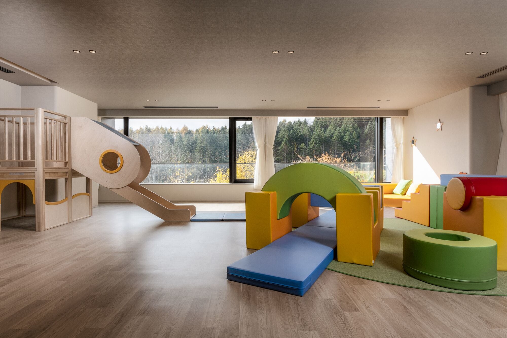 A spacious room with a wooden play area, large windows, and a view of trees.