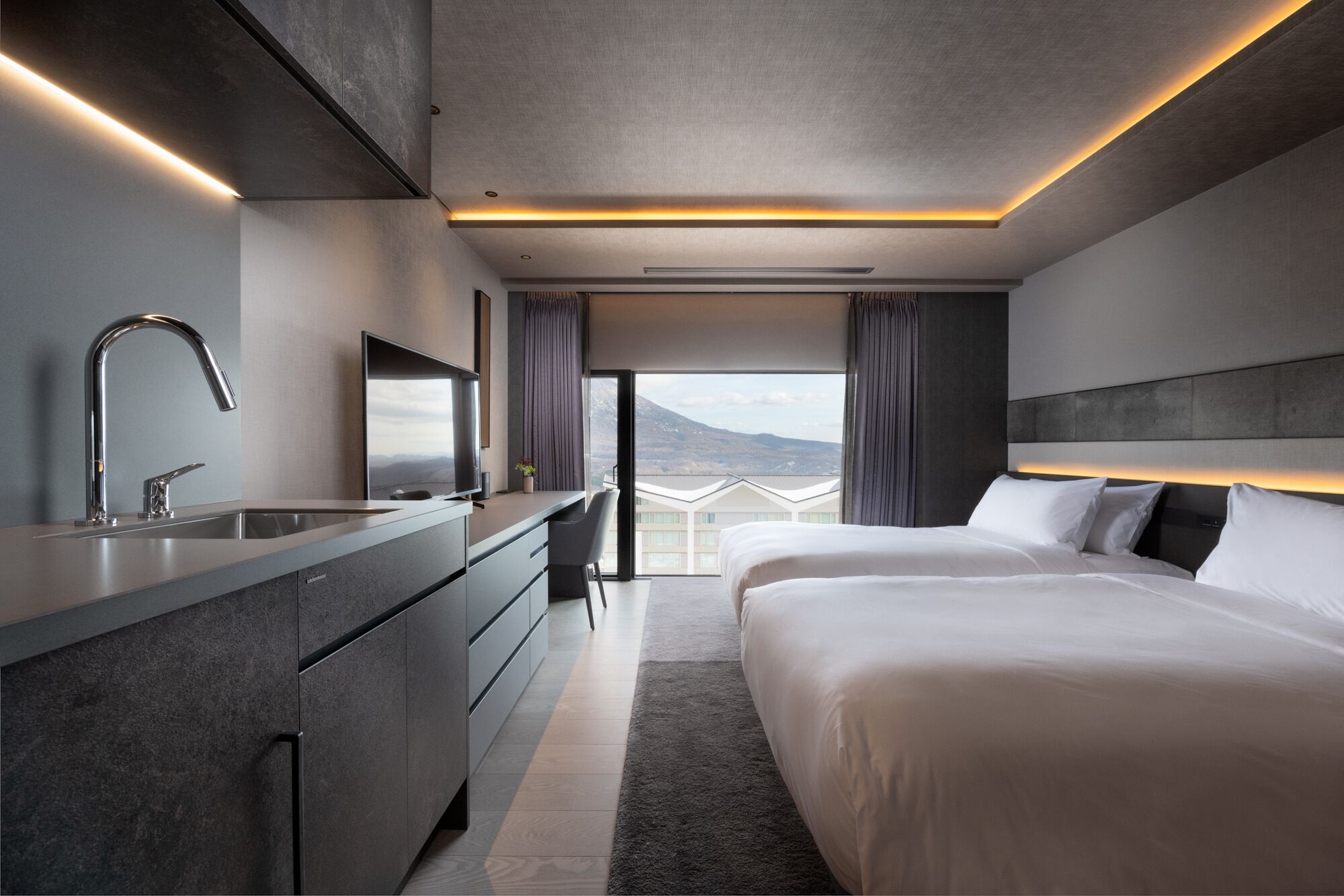 A modern hotel room with a large bed, a built-in desk, a flat-screen TV, and a view of mountains through large windows.