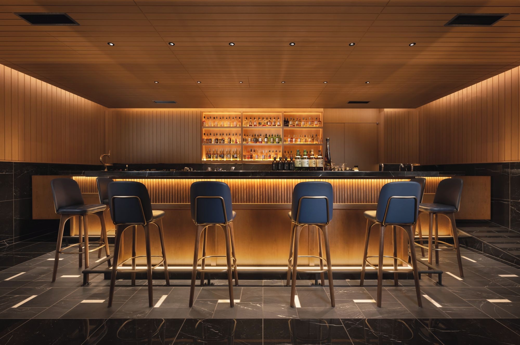 A bar area with a long counter, multiple stools, and a well-stocked shelf of bottles.