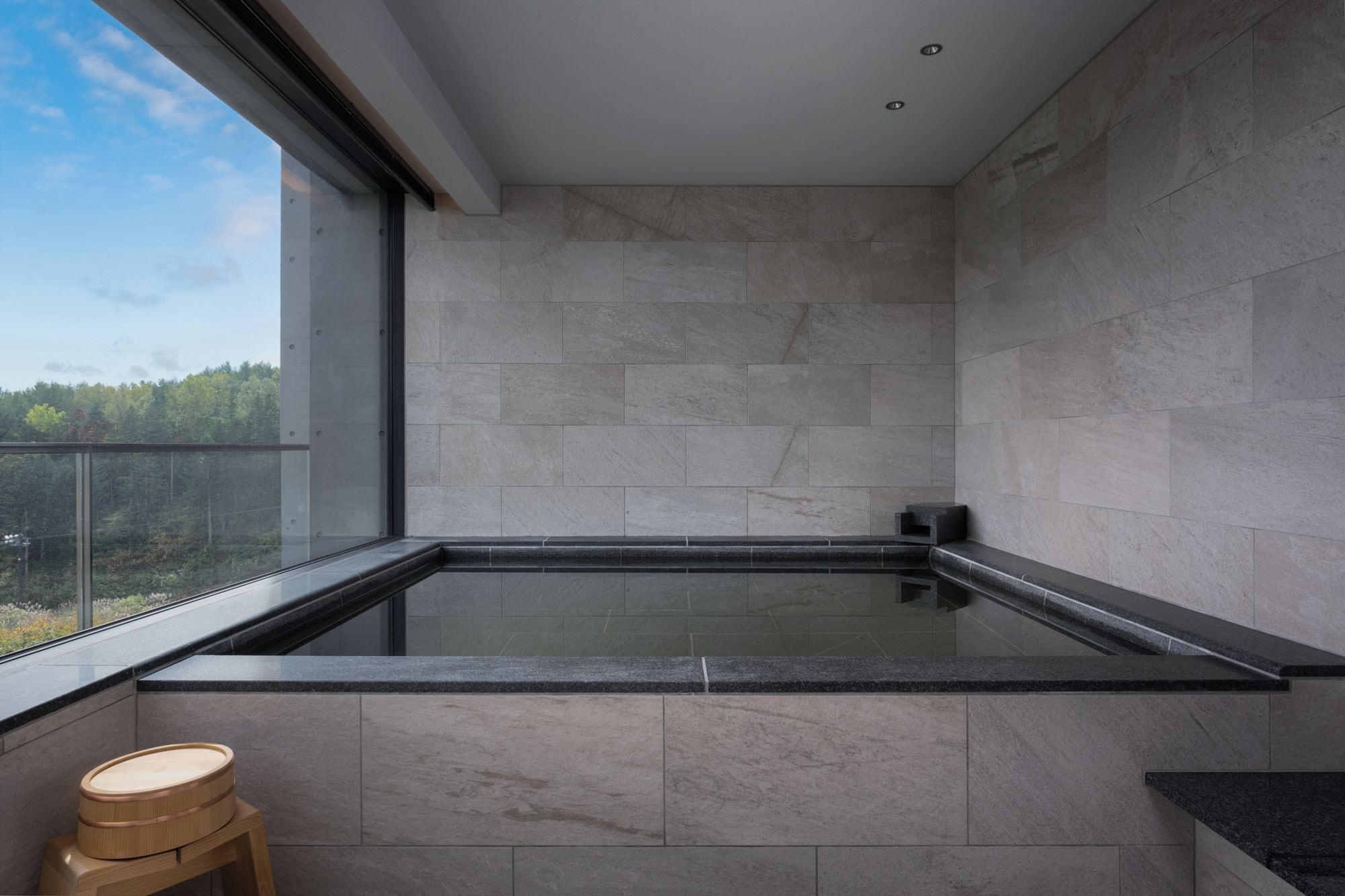 A modern bathroom with a glass-enclosed hot tub, a wooden stool, and a view of a forested area.