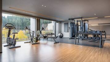 Un gimnasio moderno con máquinas de cardio, pesas libres y un amplio ventanal con vistas a árboles y un edificio.