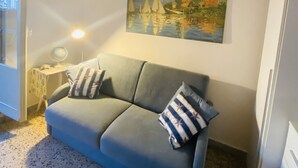 Una habitación con un sofá azul, un cuadro de veleros en la pared, un escritorio con lámpara y un equipo de aire acondicionado.
