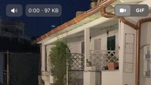 Un camino conduce a un edificio blanco con porche cubierto, plantas en macetas y una ventana visible.