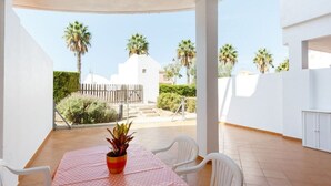 Eine überdachte Terrasse im Freien mit Tisch und Stühlen, einem Blumentopf und Blick auf Palmen sowie ein weißes Gebäude im Hintergrund.