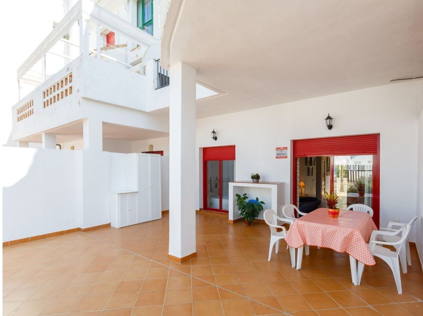 A spacious outdoor area with a dining table and chairs, a red-framed door, and a balcony with white railings.