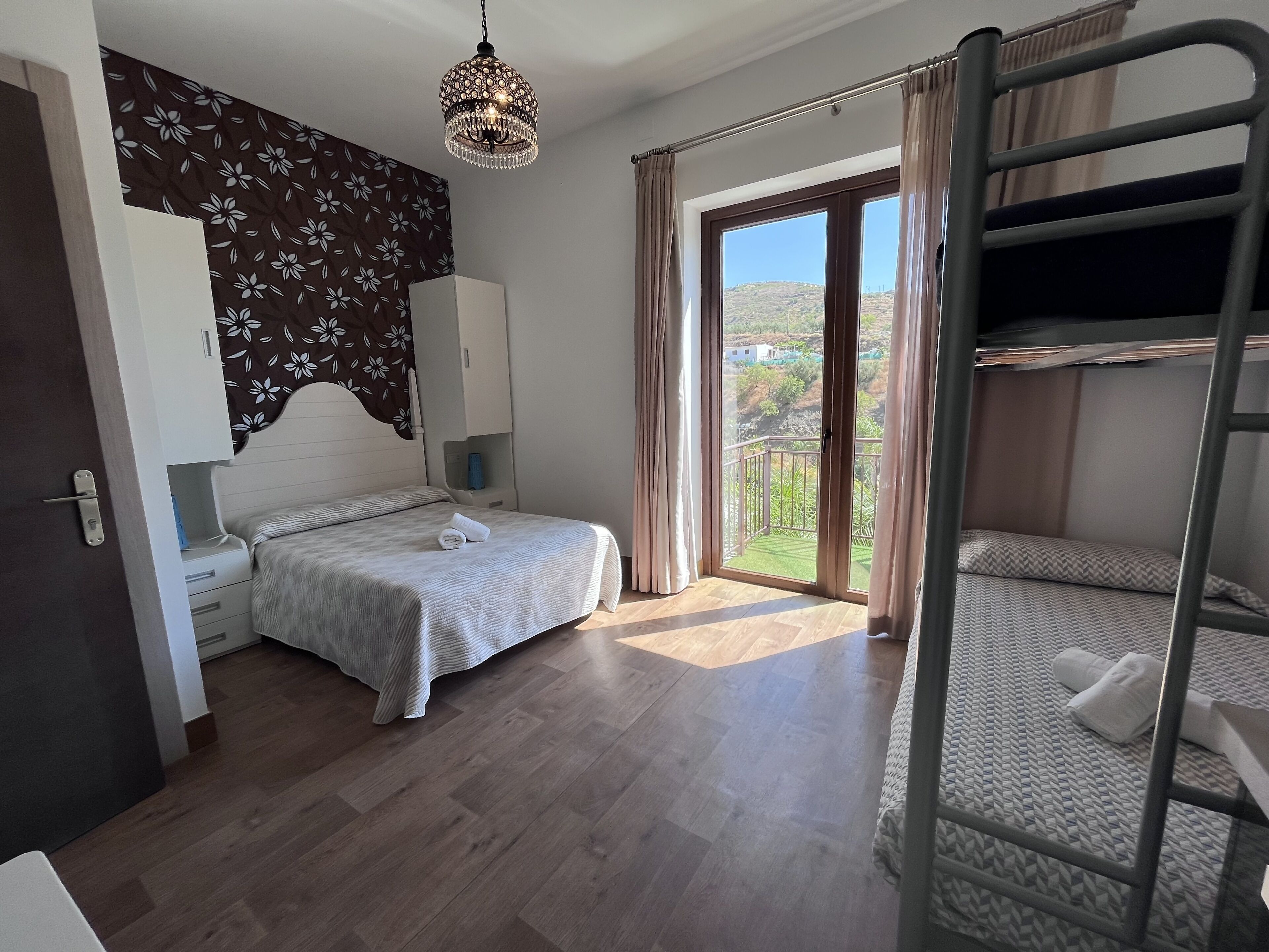 A bedroom with a bunk bed, a single bed, a wardrobe, a bedside table, a hanging light fixture, and a view of a green landscape through a sliding door.