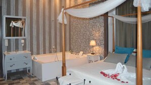 A four-poster canopy bed with a white canopy, a large bathtub, a vanity with a mirror, and a nightstand with a lamp. - Suite with private jacuzzi in the room (Arenas)