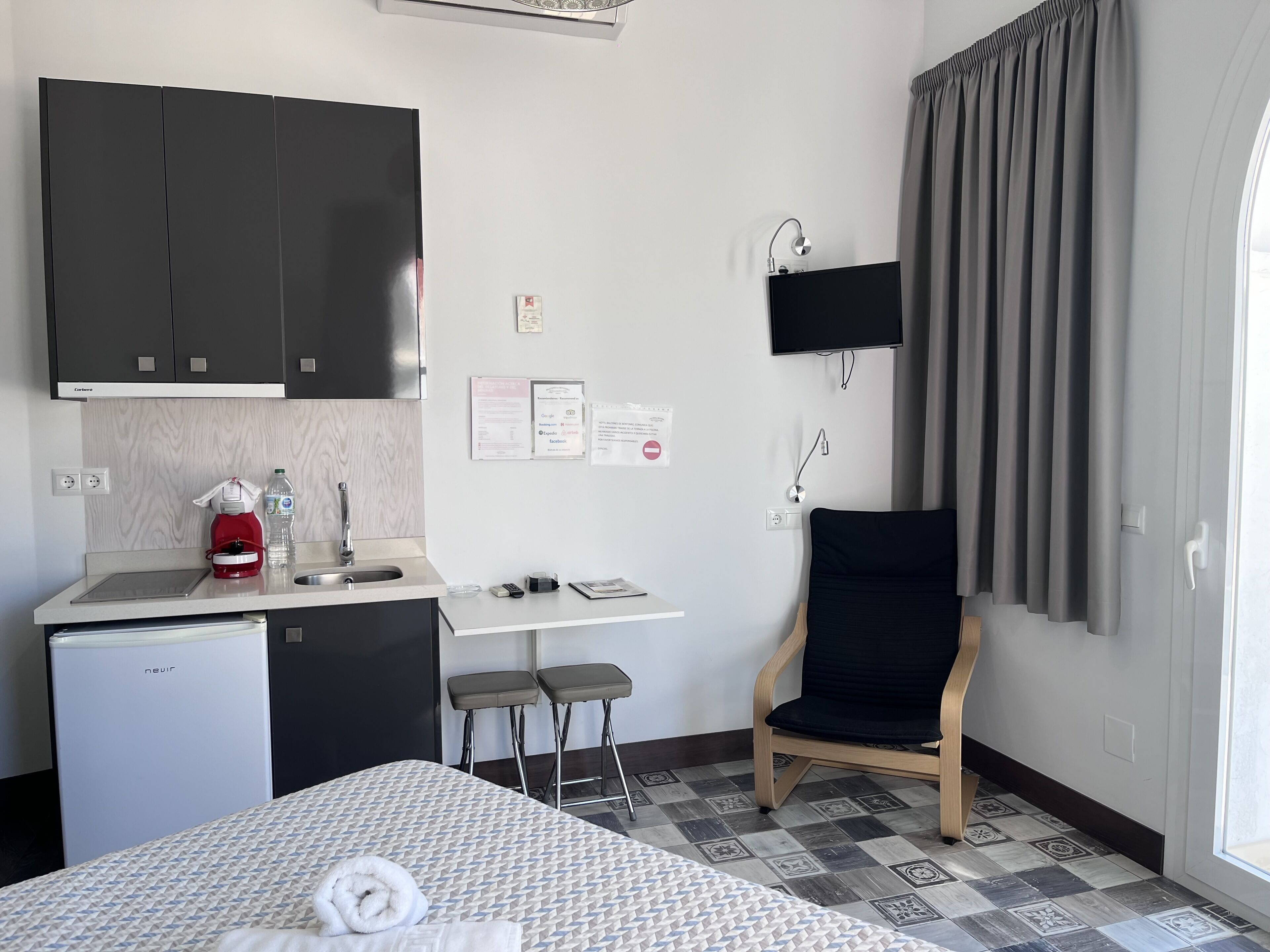 A compact hotel room with a kitchenette, a bed with a patterned bedspread, a black chair, and a window with curtains.