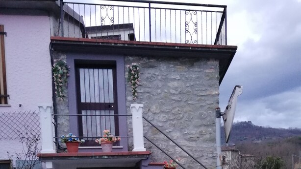 Una casa a due piani con facciata in pietra, una porta d'ingresso scura e un piccolo balcone con piante in vaso.