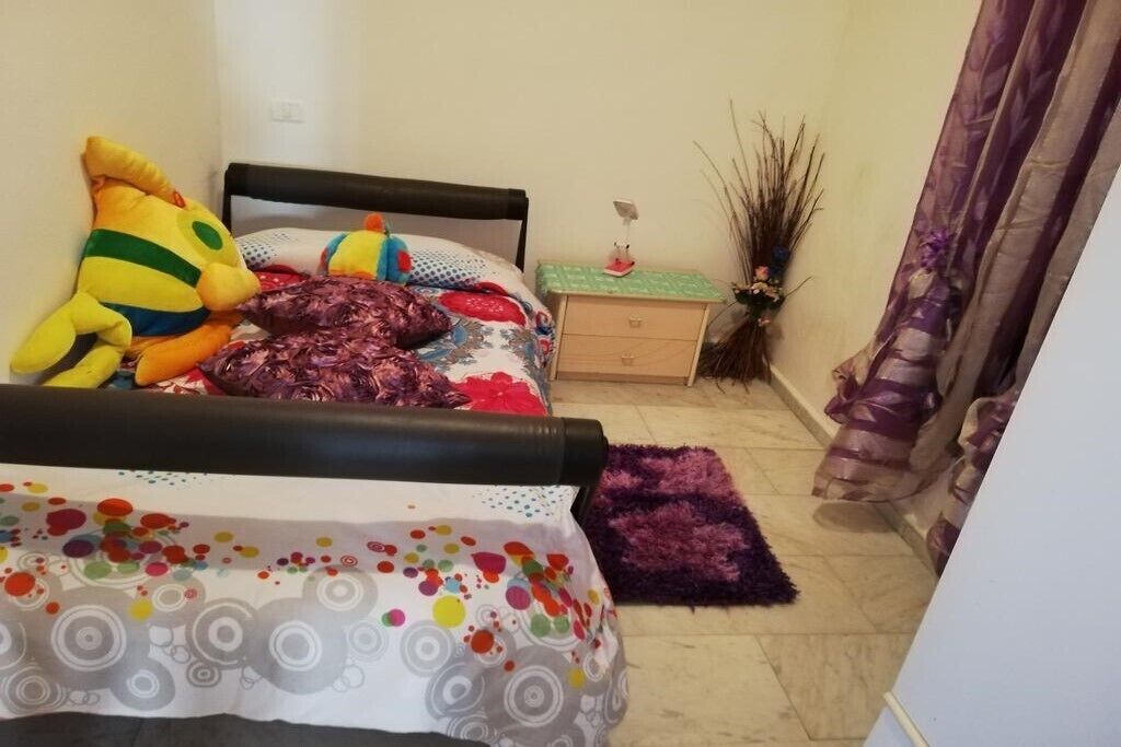 Ein Schlafzimmer mit einem Bett, einem Nachttisch, einer Kommode, einem violetten Teppich und einer Zierpflanze.