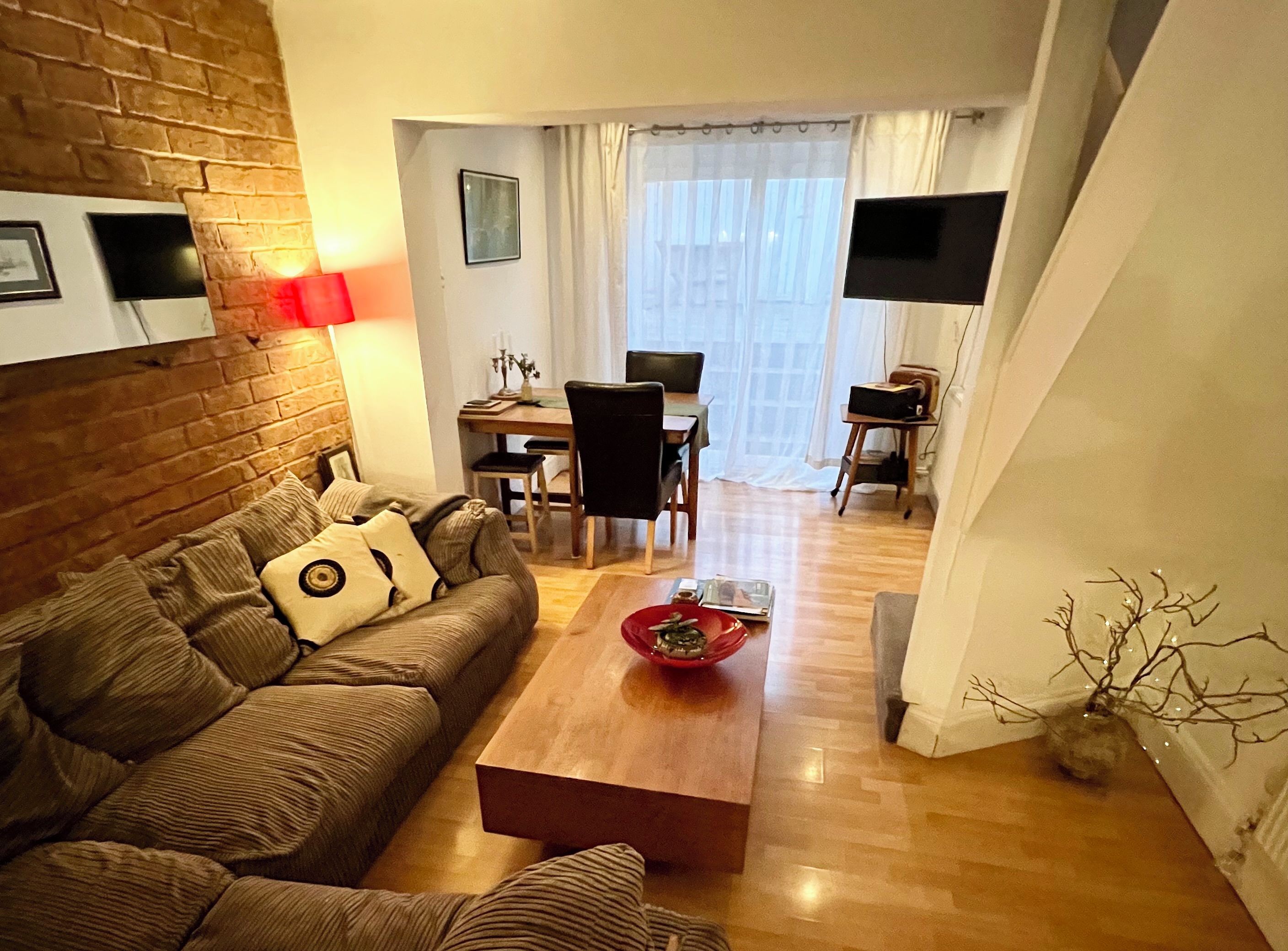 A cozy living room with a brick wall, a wooden coffee table, a sofa with cushions, a dining table with chairs, a flat-screen TV, and a window with curtains.