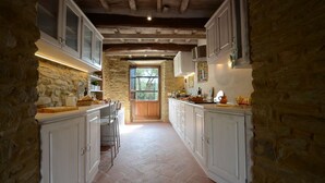 A narrow stone-walled kitchen with white cabinetry, a wooden ceiling, and a window providing natural light. - Torre La Ripa 8, Emma Villas (Cortona)