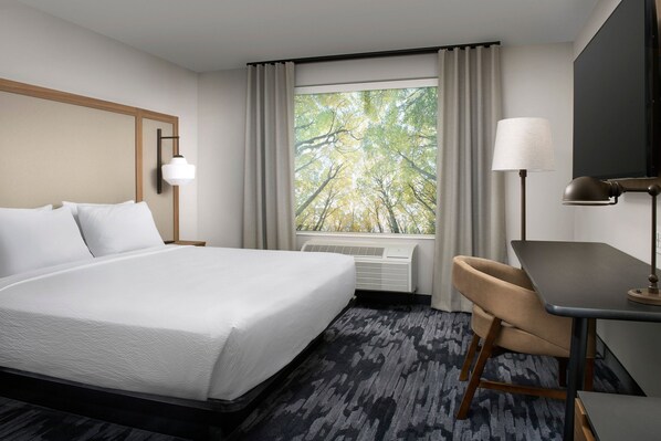 A hotel room with a large bed, a desk, a chair, a window with curtains, and a wall-mounted lamp.