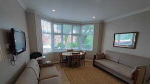 Ein Wohnzimmer mit einer Couch, einem Esstisch mit Stühlen, einem an der Wand montierten Fernseher und großen Fenstern mit Jalousien.