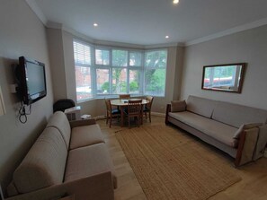 A living room with a sofa, a dining table with chairs, a TV mounted on the wall, and large windows with blinds.