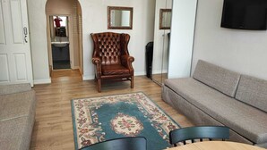 Ein Wohnzimmer mit einer Couch, einem Holzesstisch, einem gepolsterten Ledersessel, einem gemusterten Teppich und einem an der Wand befestigten Fernseher.
