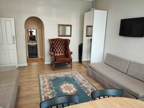 Ein Wohnzimmer mit einer Couch, einem Holzesstisch, einem gepolsterten Ledersessel, einem gemusterten Teppich und einem an der Wand befestigten Fernseher.