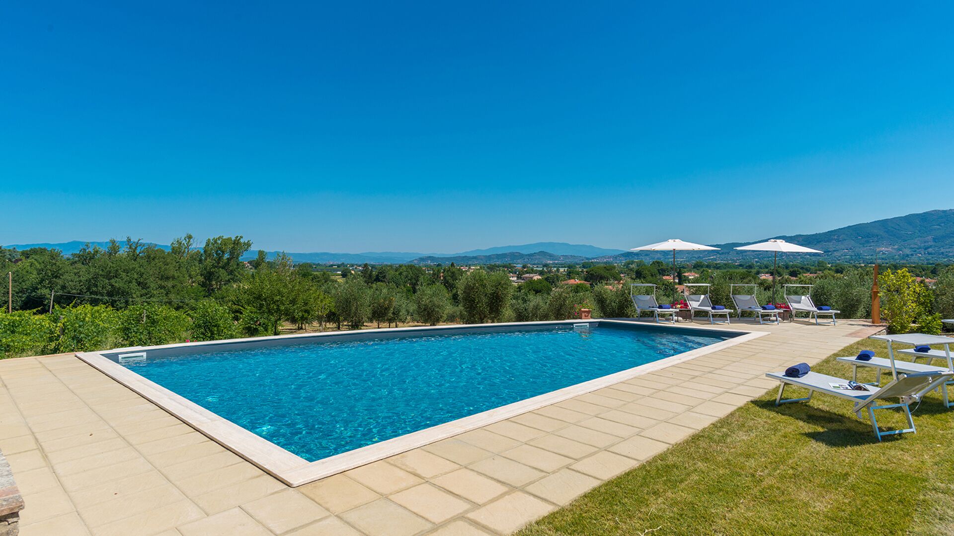 A rectangular swimming pool with a tiled deck, surrounded by lounge chairs and umbrellas, with a view of a mountainous landscape.