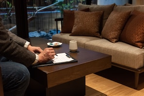 A person sitting at a wooden table with a clipboard, a plate of food, and a cup. In the background, there is a beige sofa with brown cushions.