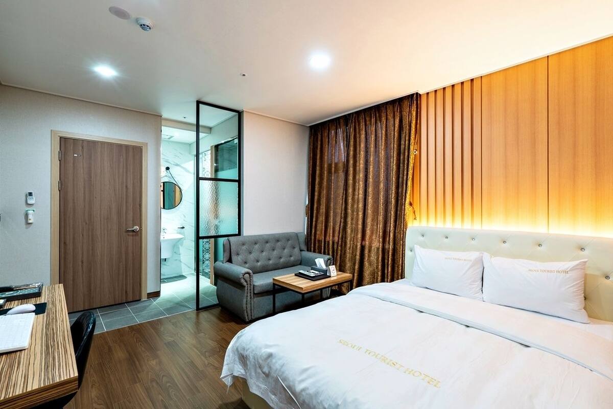 Habitación de hotel con cama, escritorio, silla, armario y baño visible a través de una mampara de vidrio.