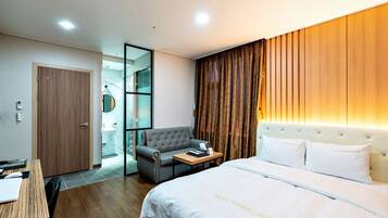 A hotel room with a bed, a desk, a chair, a wardrobe, and a bathroom visible through a glass partition.