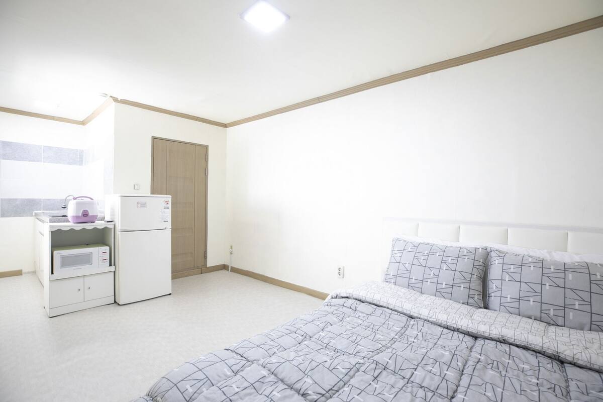 A bedroom with a bed, a small kitchenette with a refrigerator and microwave, and a wooden door.