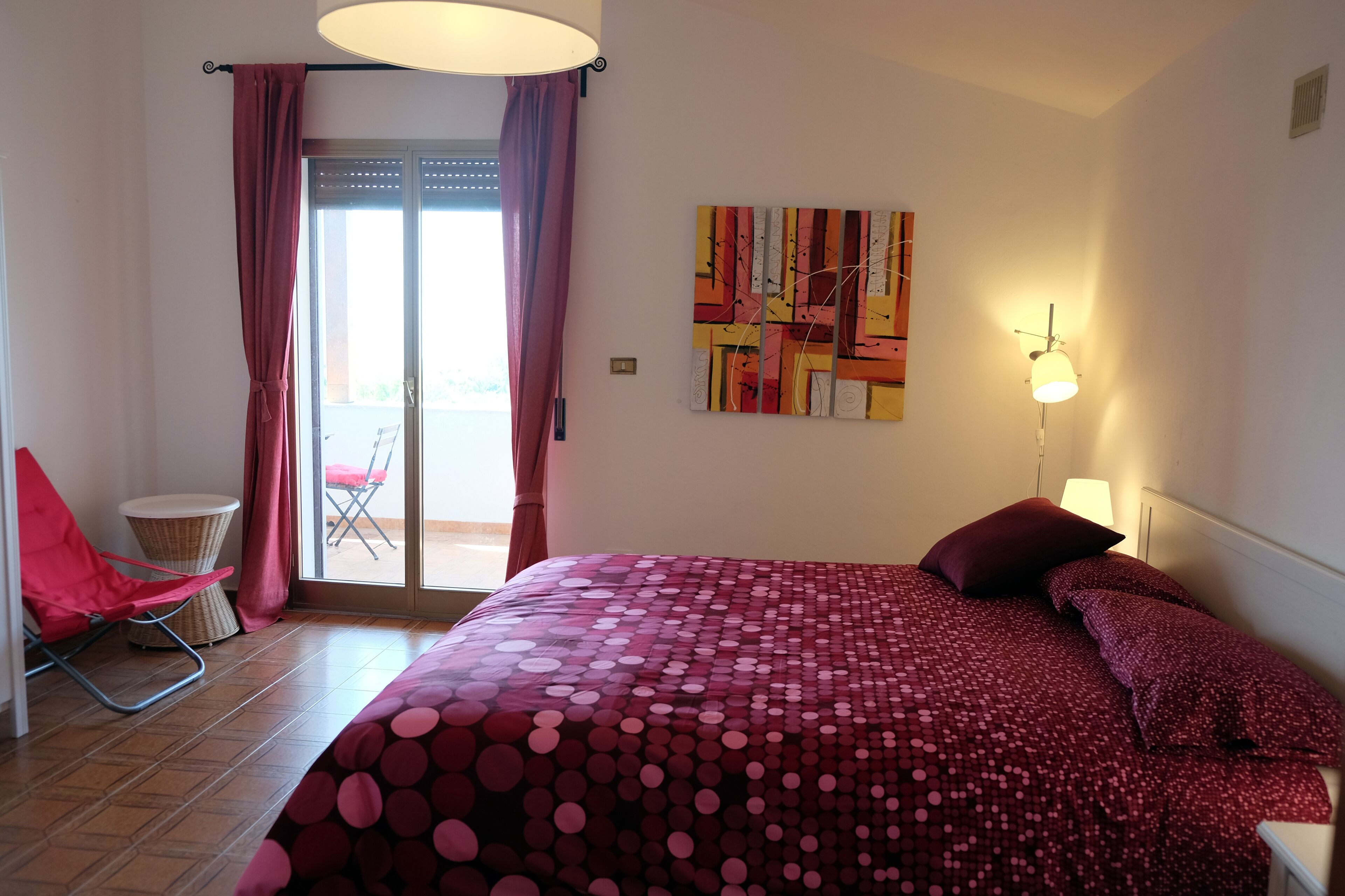 A bedroom with a large bed, a chair, a lamp, and a view of the outdoors.