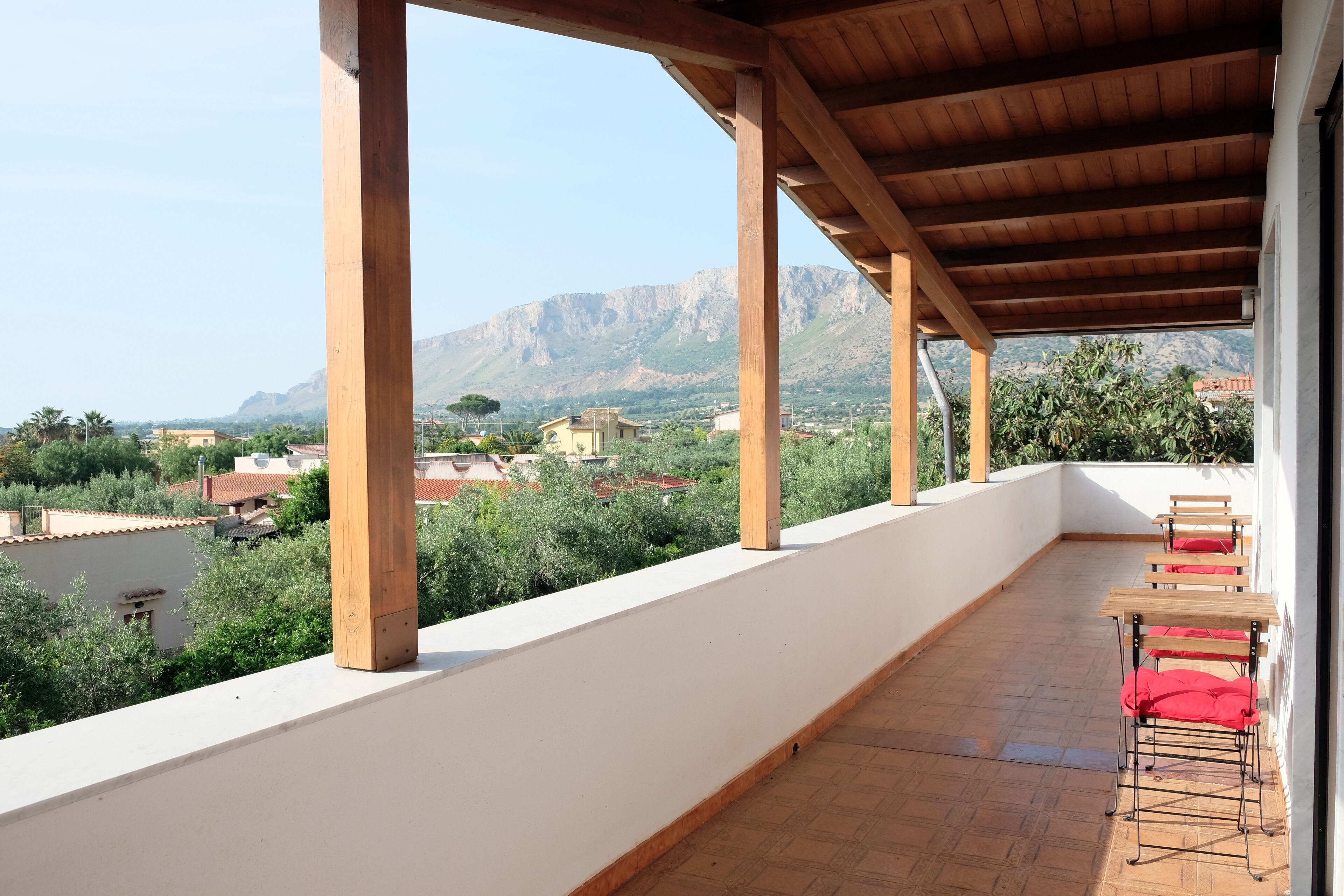 Un balcón con una pérgola de madera, una mesa con cojines rojos, y vistas a montañas y edificios.