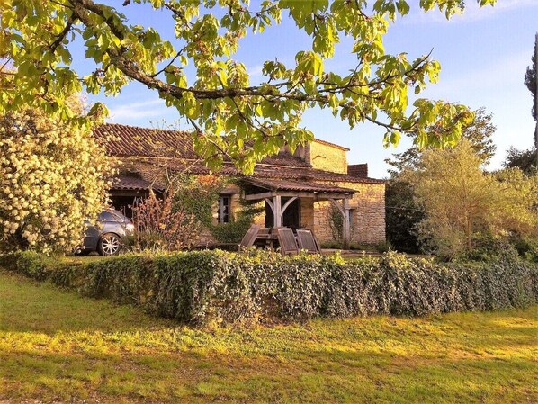 A rustic stone house with a tiled roof, surrounded by a well-maintained hedge and a car parked in front.