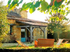 A stone house with a tiled roof, a covered patio with wooden furniture, and a view of the surrounding garden.
