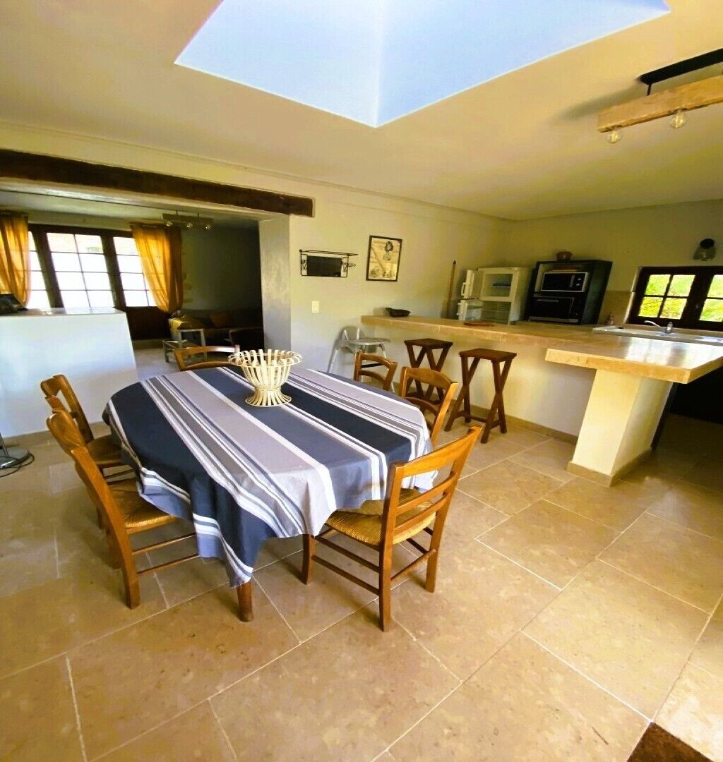 Un coin repas avec une table dressée pour six personnes, des chaises en bois, un espace cuisine équipé d’appareils électroménagers et une lucarne au-dessus.