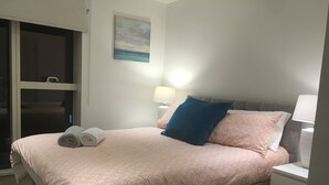 Una cama bien hecha con una colcha estampada en tonos rosa, dos almohadas (una azul y otra rosa), y dos toallas enrolladas. Se ve una mesita de noche con una lámpara, un cuadro en la pared y una vista a través de una puerta de cristal.