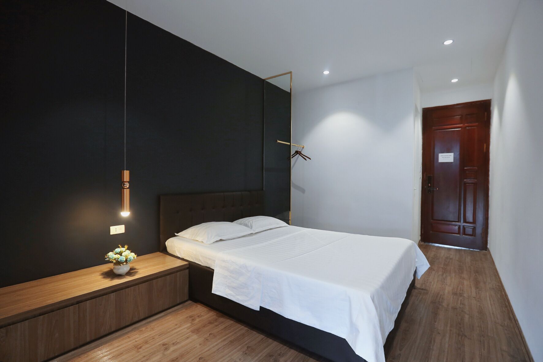 A modern bedroom with a large bed, a bedside table with a vase, a mirror, and a wooden floor.