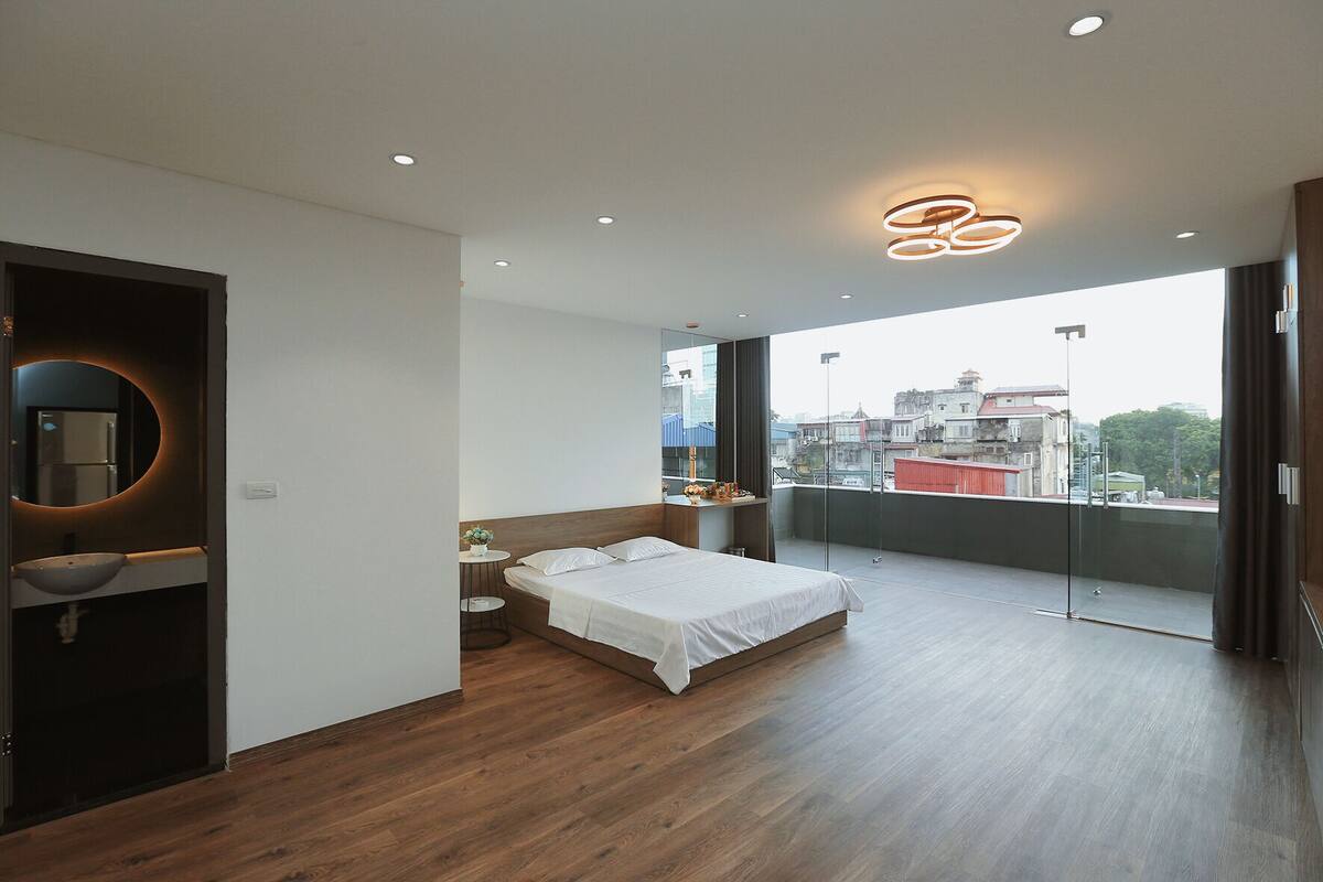 A modern bedroom with a large bed, a round mirror, a small table, and a view of the city through large windows.