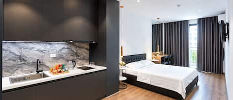 A modern hotel room with a kitchen area, a bed with a dark headboard, and a view of the outdoors through large windows.