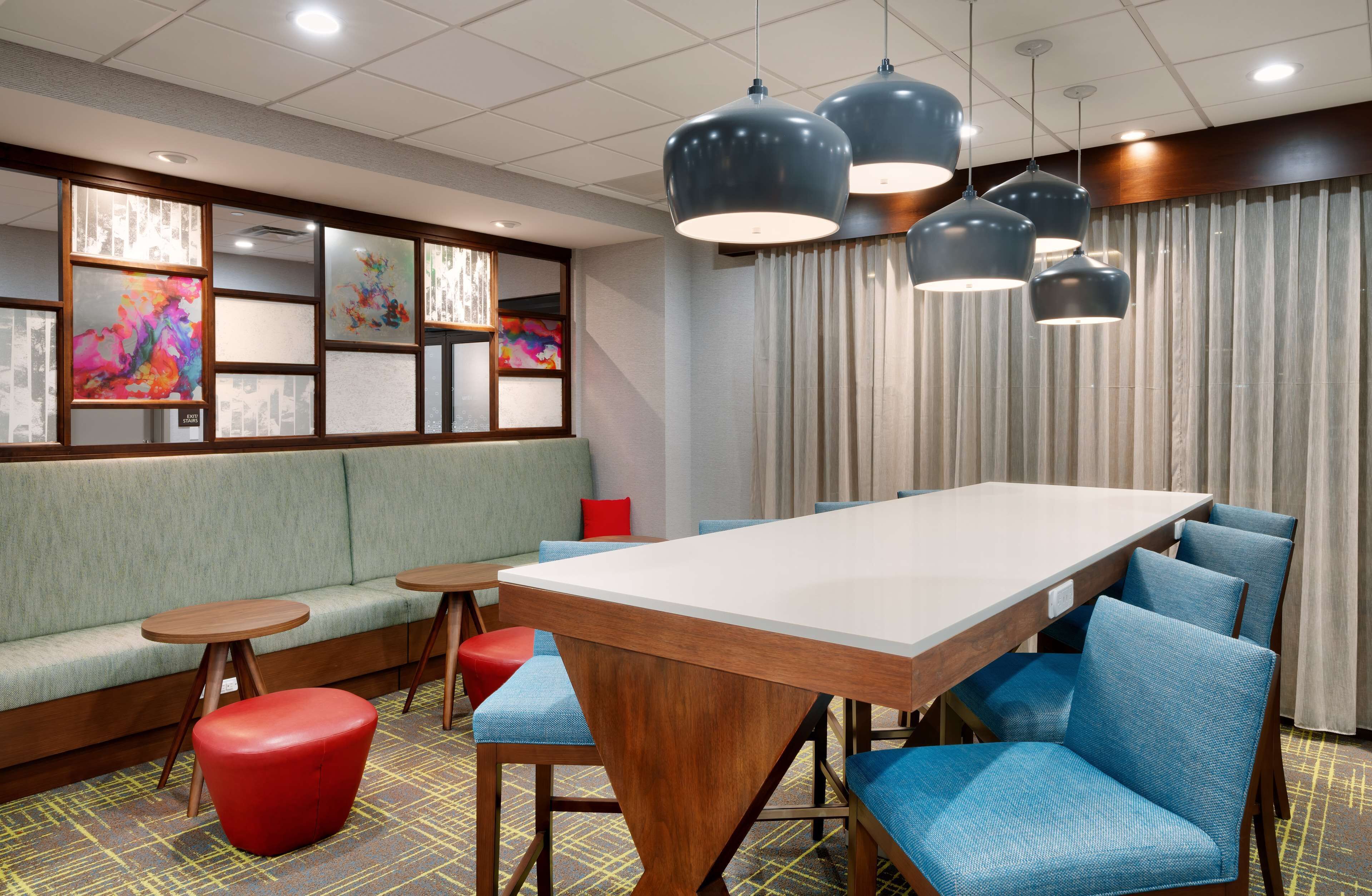 Un hall d’hôtel moderne avec une longue table, des chaises à revêtement bleu, un pouf rouge et un canapé vert.