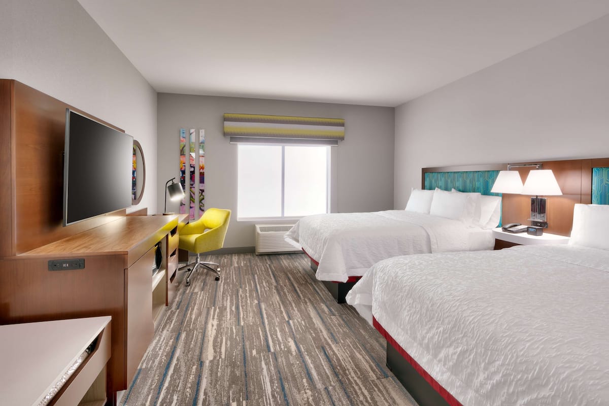A hotel room with a flat-screen TV, a desk, a yellow chair, two beds, and a window with a view.
