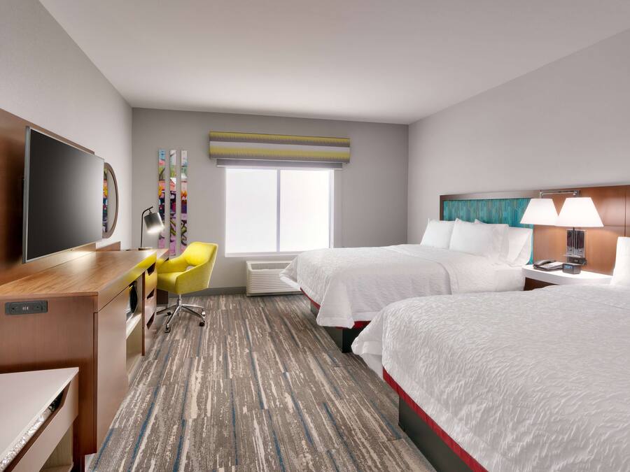 A hotel room with a flat-screen TV, a desk, a yellow chair, two beds, and a window with a view.