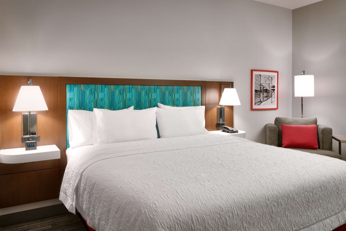 A hotel room with a large bed, two bedside tables with lamps, a headboard with a geometric pattern, a red pillow, and a framed picture on the wall.