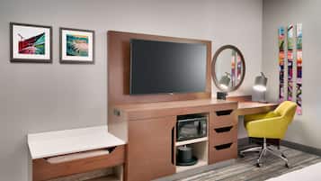 A modern hotel room with a wooden desk, a flat-screen TV, a mirror, and a yellow chair. There are two framed artworks on the wall.