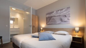 Un dormitorio moderno con una cama grande, mesitas de noche, un armario de madera, un baño con espejo y lavamanos, y un cuadro colgado en la pared.
