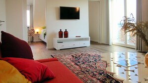 Ein modernes Wohnzimmer mit einem an der Wand befestigten Flachbildfernseher, einem weißen Konsolentisch mit zwei Flaschen, einem gemusterten Teppich und einem roten Sofa.