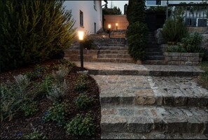 Un escalier en pierre mène à un bâtiment blanc, entouré d’une clôture en bois et agrémenté d’un jardin où poussent différentes espèces de plantes.