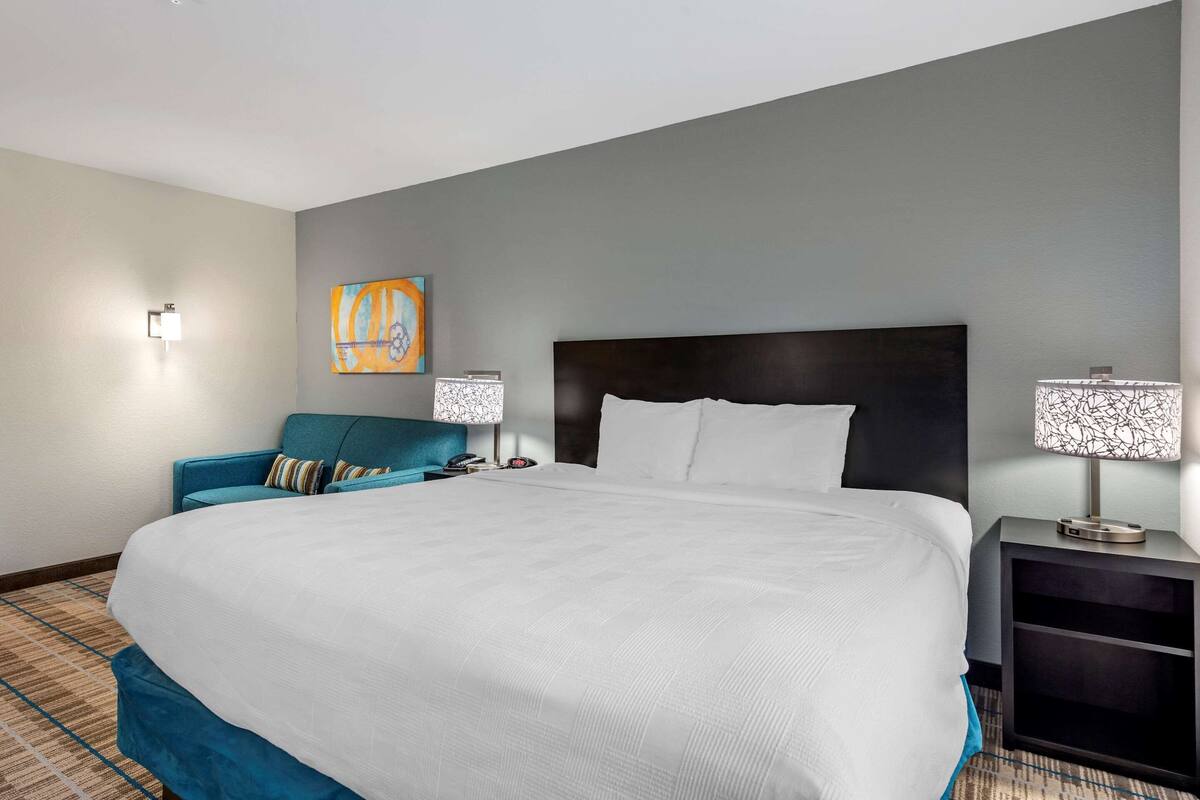 Een hotelkamer met een groot bed, een blauwe bank, twee nachtkastjes met lampen, en een kunstwerk aan de muur.
