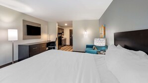 A modern hotel room with a large bed, a desk, a flat-screen TV, a blue sofa, and a kitchenette in the background.