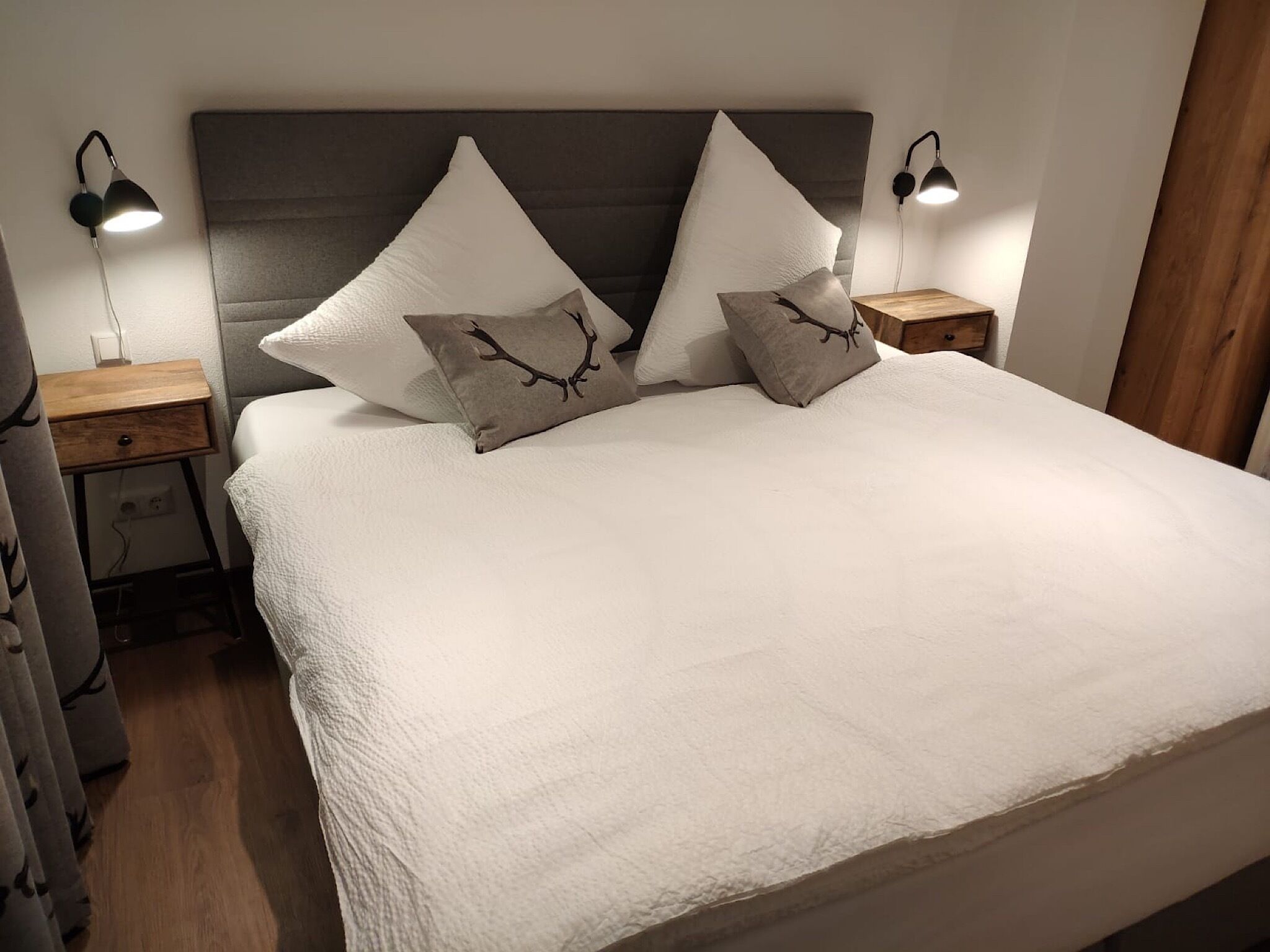 Een net opgemaakt bed met een witte deken en twee decoratieve kussens, aan weerszijden geflankeerd door twee nachtkastjes met lampen, in een eenvoudige, moderne kamer.